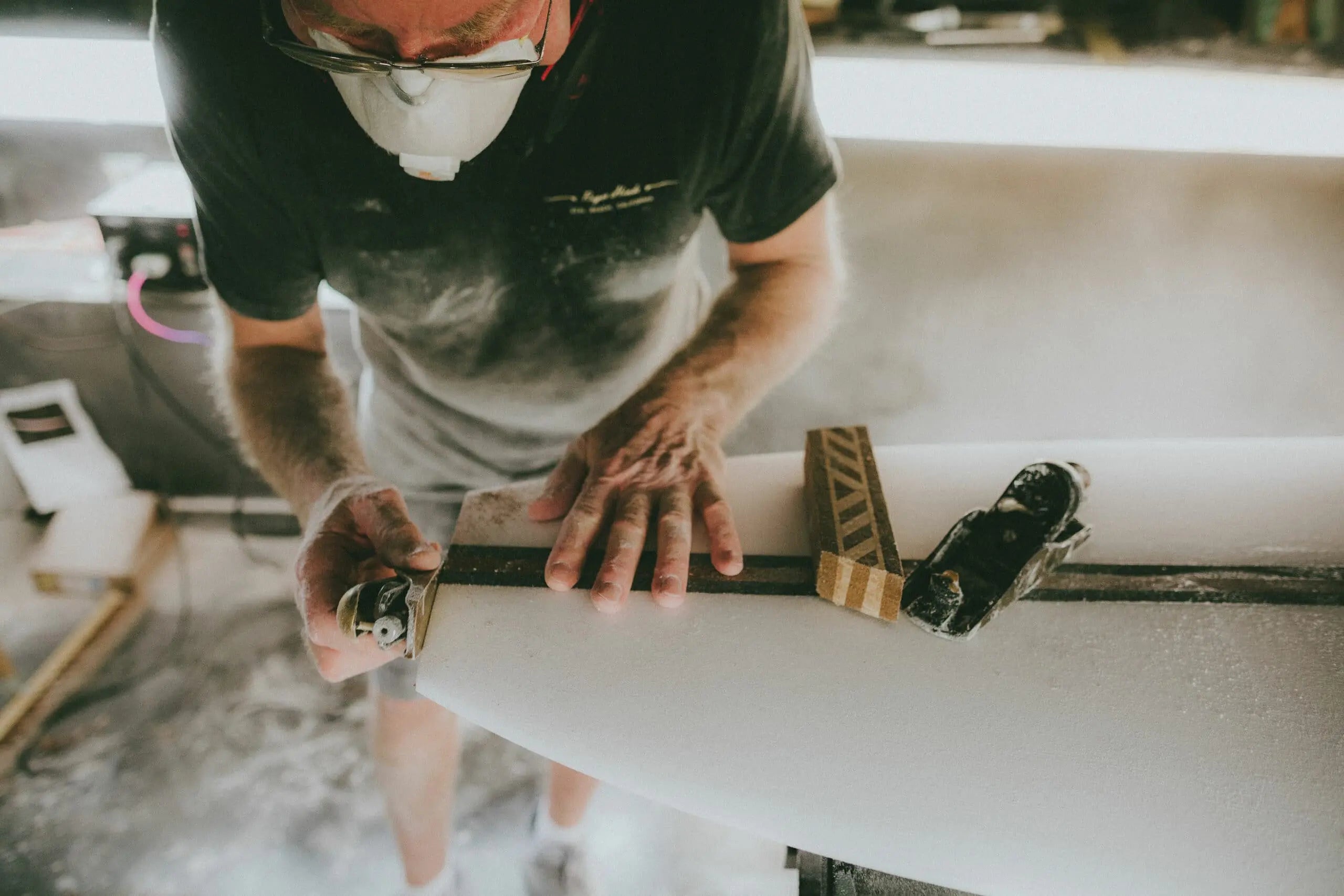 Shapers-Club- Un artisan du surf portant un masque anti-poussière et des lunettes de protection façonne une planche de surf avec un outil de rabotage, créant ainsi de la poussière de mousse dans un environnement d'atelier. -surfshop-surfboard