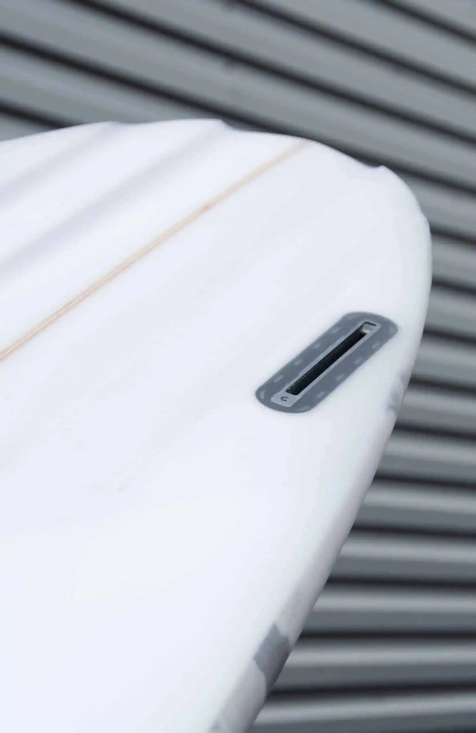 Shapers-Club- Gros plan de la queue d'une planche de surf blanche Aphex 6'10" x 20' 3/4" x 2' 3/4", mettant en valeur son boîtier à aileron unique, avec un revêtement en métal ondulé gris en arrière-plan. -surfshop-surfboard