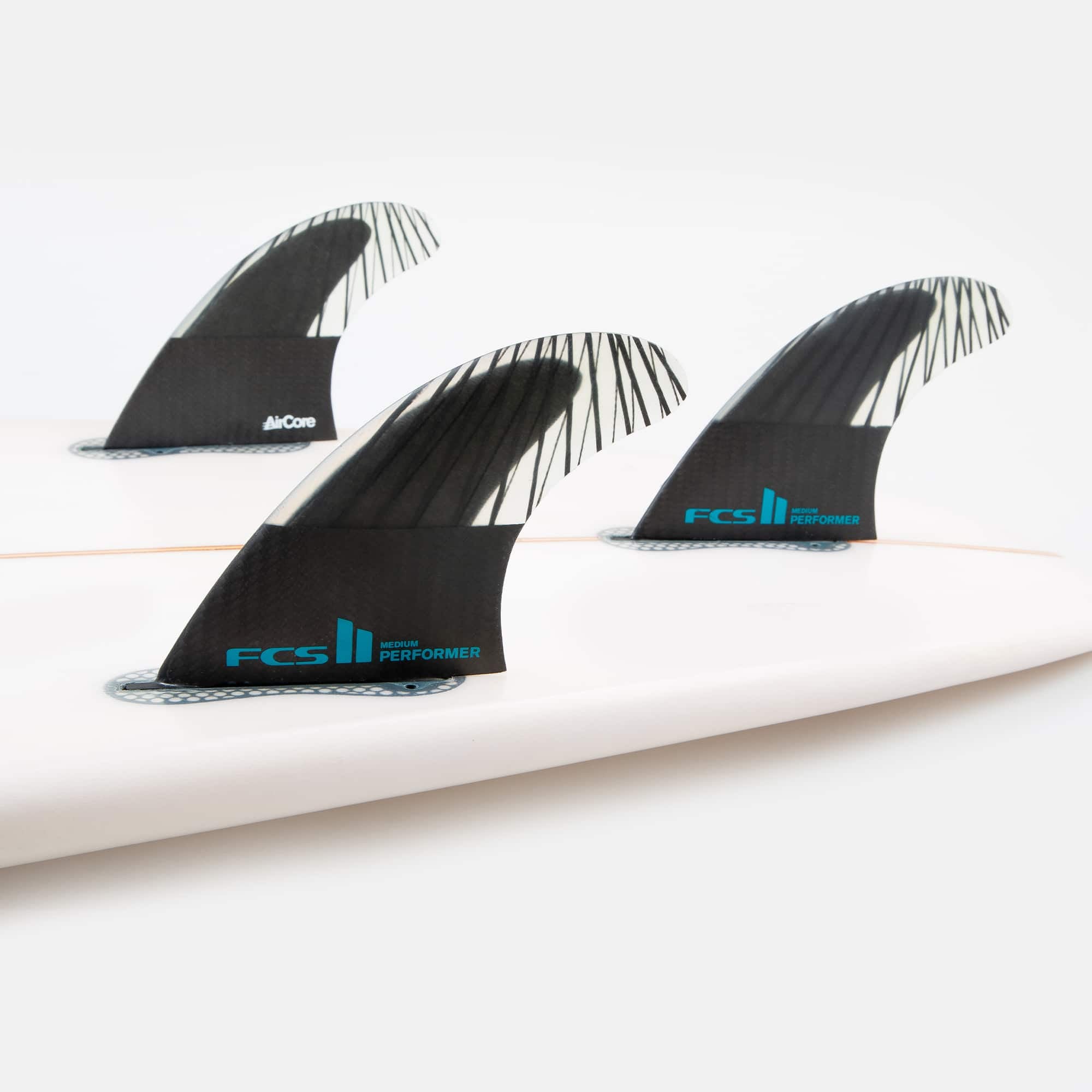 Shapers-Club- Image en gros plan d'une planche de surf blanche équipée de trois ailerons noirs FCS II - PERFORMANCE - THRUSTER (Performance Core Carbon) avec un logo bleu et le texte « Performer » sur chaque aileron, disposés selon une disposition de type thruster axée sur la performance. Le fond est blanc uni. -surfshop-surfboard