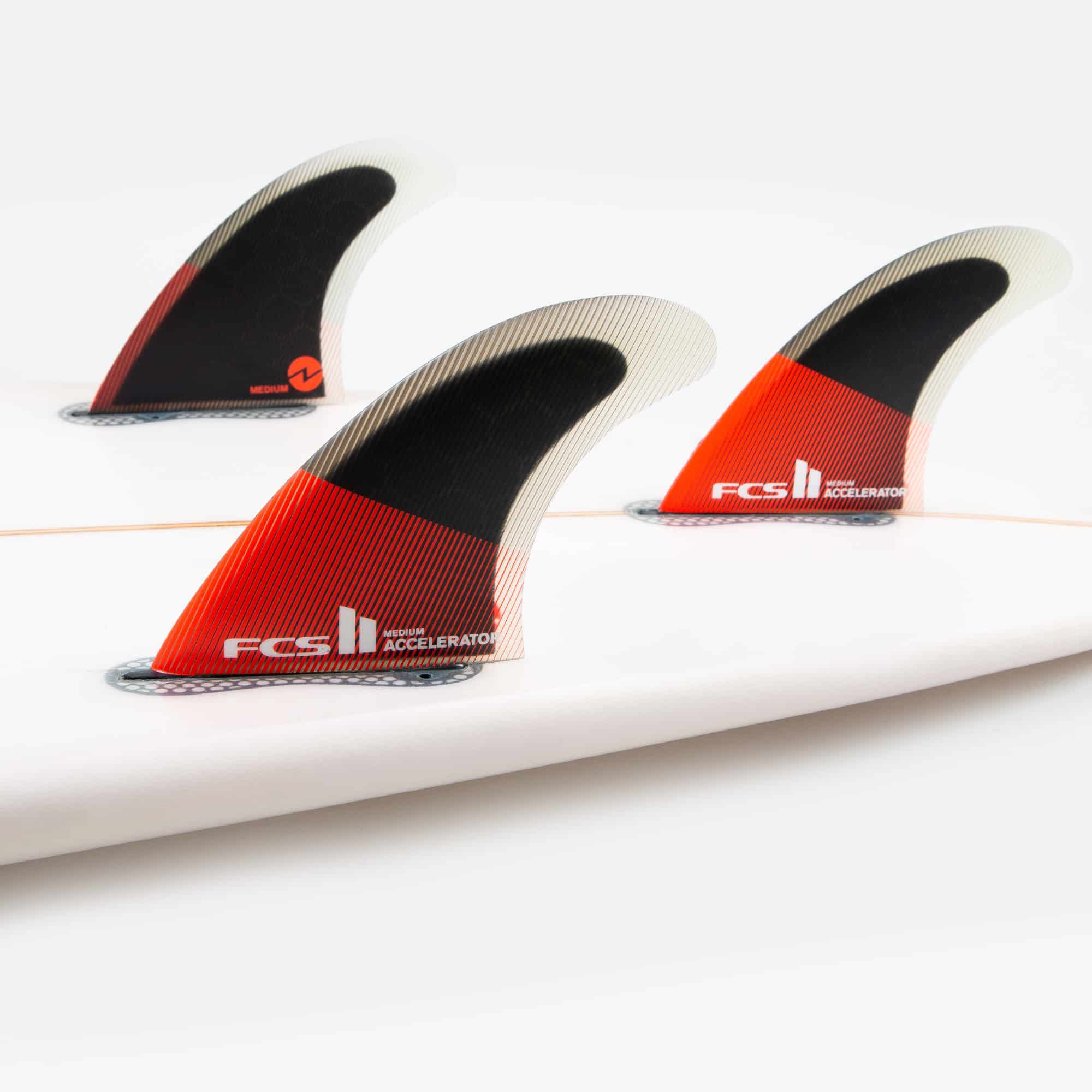Shapers-Club- Gros plan de trois ailerons FCS II - ACCELERATOR - THRUSTER (Performance Core) fixés à l'arrière d'une planche de surf. Les ailerons noirs, rouges et blancs comportent le texte « FCS II ACCELERATOR ». La planche de surf est blanche avec un longeron orange visible qui descend au centre. -surfshop-surfboard