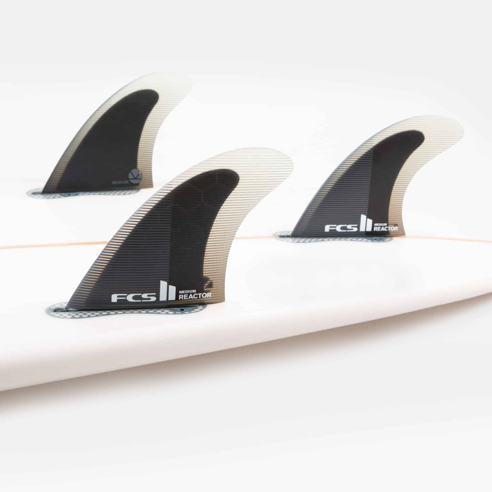Shapers-Club- Gros plan d'une planche de surf blanche dotée de trois ailerons noirs portant le logo FCS II - Reactor - Thruster (Performance Core). Les ailerons du Thruster sont profilés pour plus de performances et présentent une texture en nid d'abeille à la base. La planche de surf est placée sur un fond blanc uni. -surfshop-surfboard