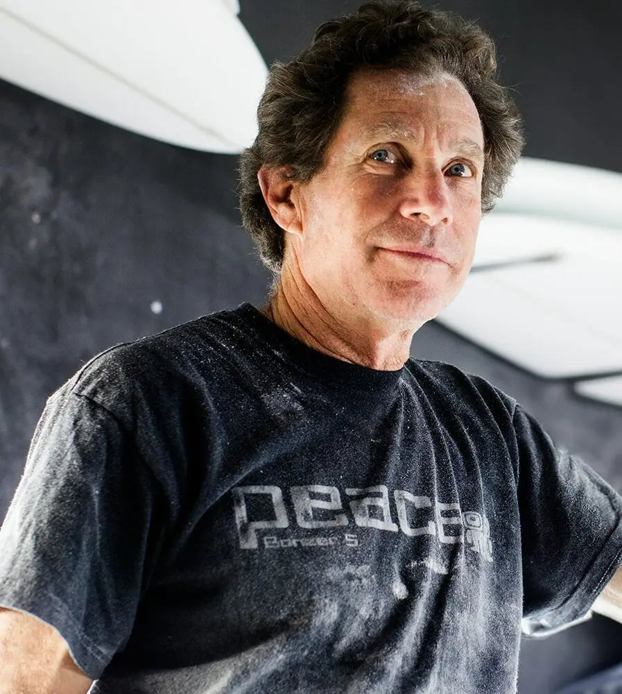 Shapers-Club- Une personne vêtue d'un T-shirt Acompte deposite - Campbell Brothers de couleur sombre avec un motif imprimé fait face à la caméra sur un fond sombre. Sa chemise semble parsemée de taches blanches, tandis que l'éclairage met en valeur son visage et le haut de son corps. -surfshop-surfboard