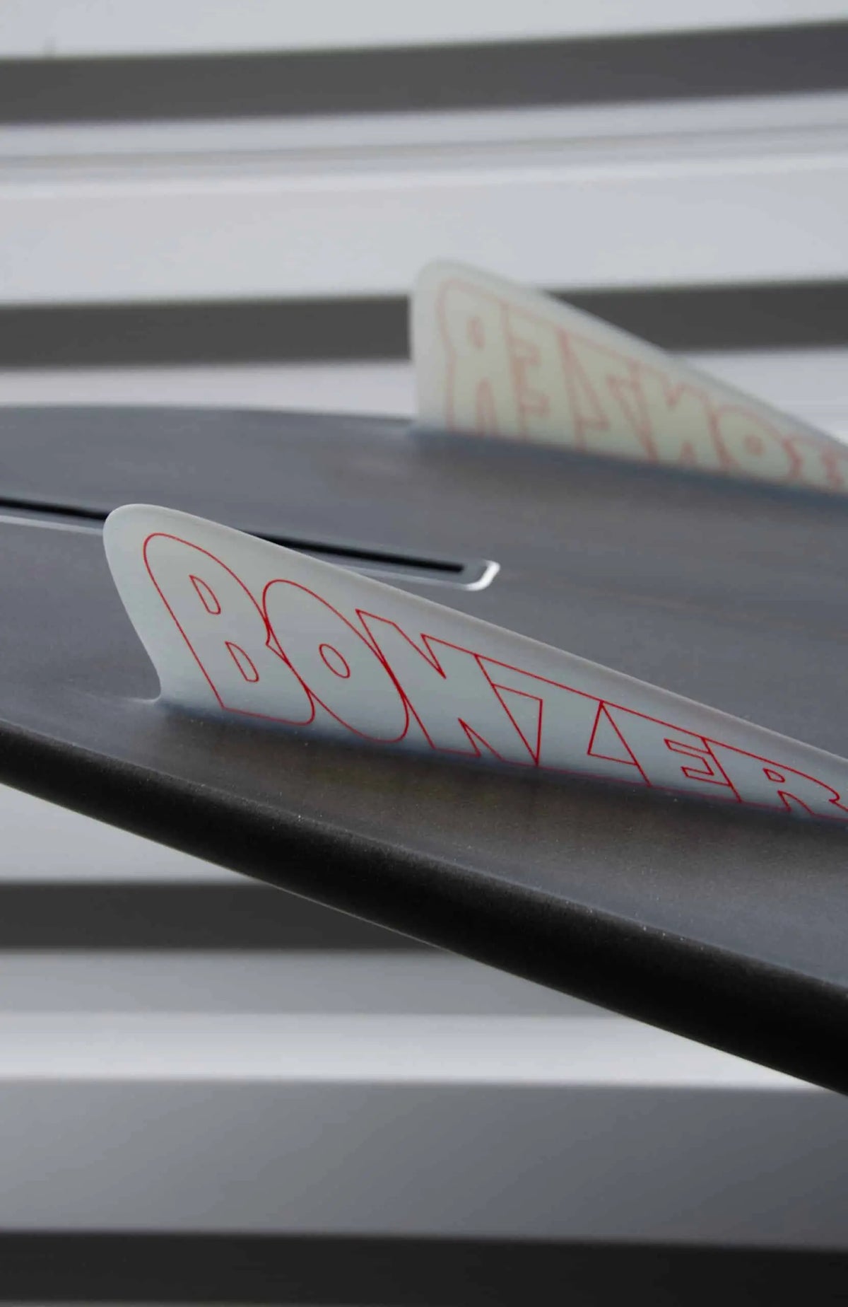 Shapers-Club- Gros plan d'un aileron de planche de surf Campbell Brothers - Russ Short avec le mot « Bonzer » écrit dessus en lettres rouges. L'arrière-plan présente des rayures horizontales floues grises et blanches. L'accent est mis sur l'aileron, offrant une vue partielle du design élégant de la planche de surf de 7'0" x 20' 3/4" x 2' 3/4". -surfshop-surfboard