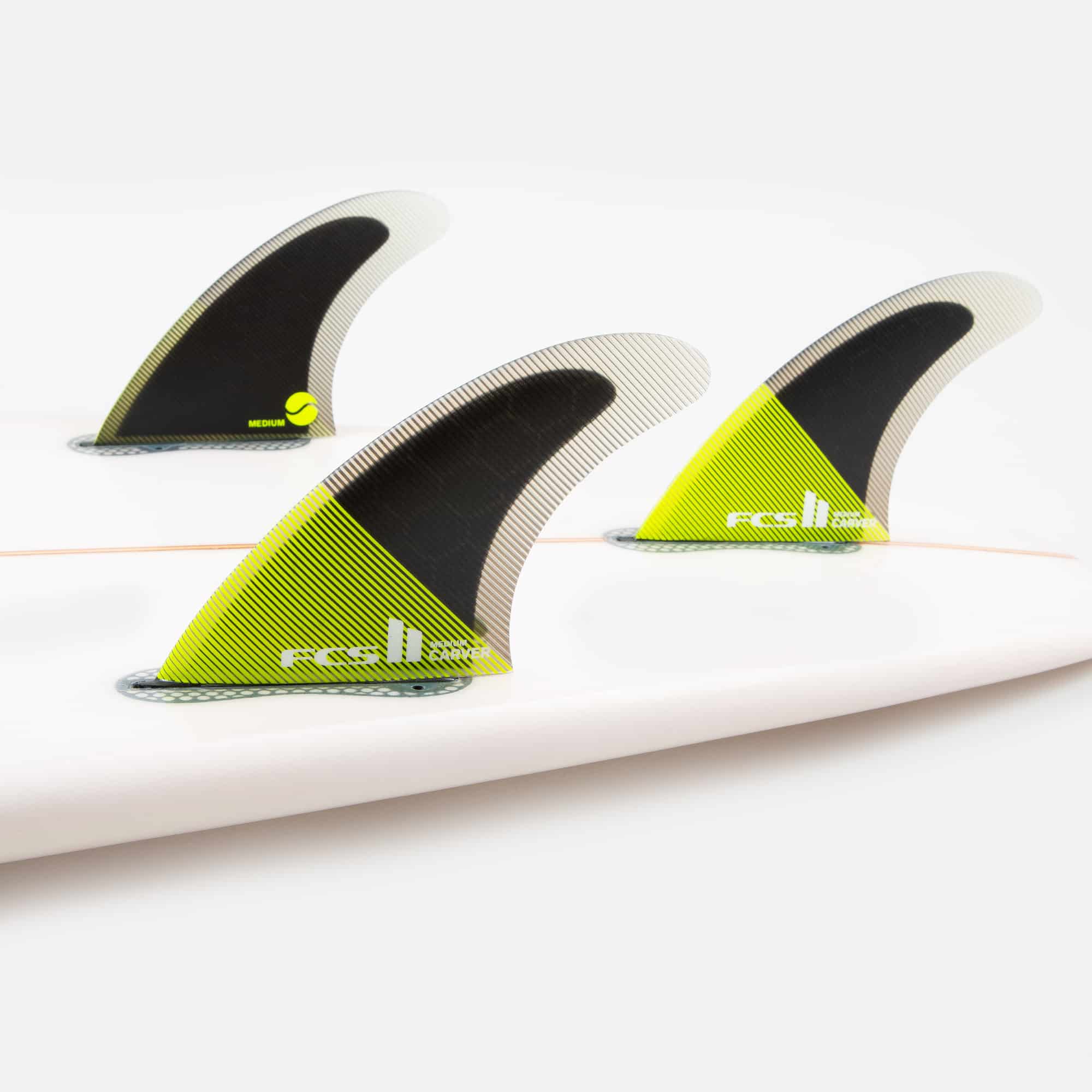 Shapers-Club- Gros plan de trois ailerons de planche de surf fixés au bas d'une planche de surf blanche. Les ailerons FCS II - CARVER - THRUSTER (Performance Core) sont principalement noirs et jaunes avec des détails texturés, étiquetés « relic ». Ces ailerons compatibles FCS II améliorent la stabilité et les performances. -surfshop-surfboard