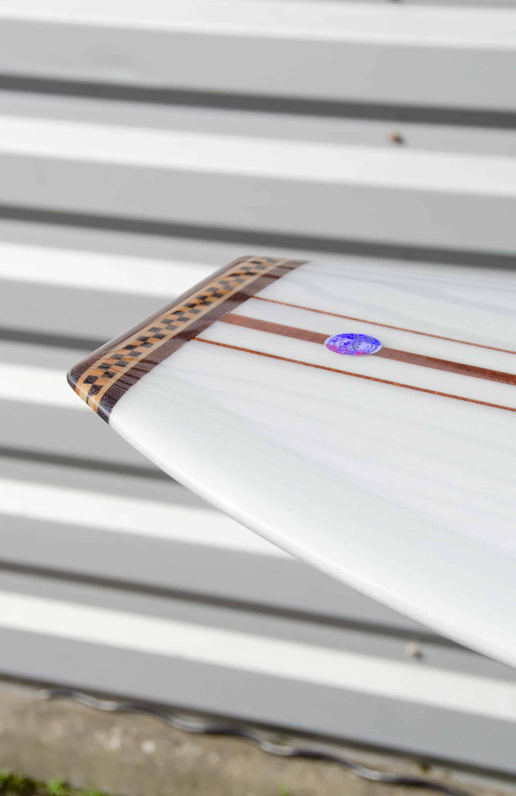 Shapers-Club- Une image en gros plan présente la queue d'une planche de surf Roger Hinds - The Classic contre un mur à rayures horizontales. Mesurant 9'6" x 23" x 3' 3/4", cette planche de surf présente un design élégant avec des incrustations de bois foncé et un petit autocollant circulaire, ajoutant une touche exquise de savoir-faire et de détail à son apparence. -surfshop-surfboard