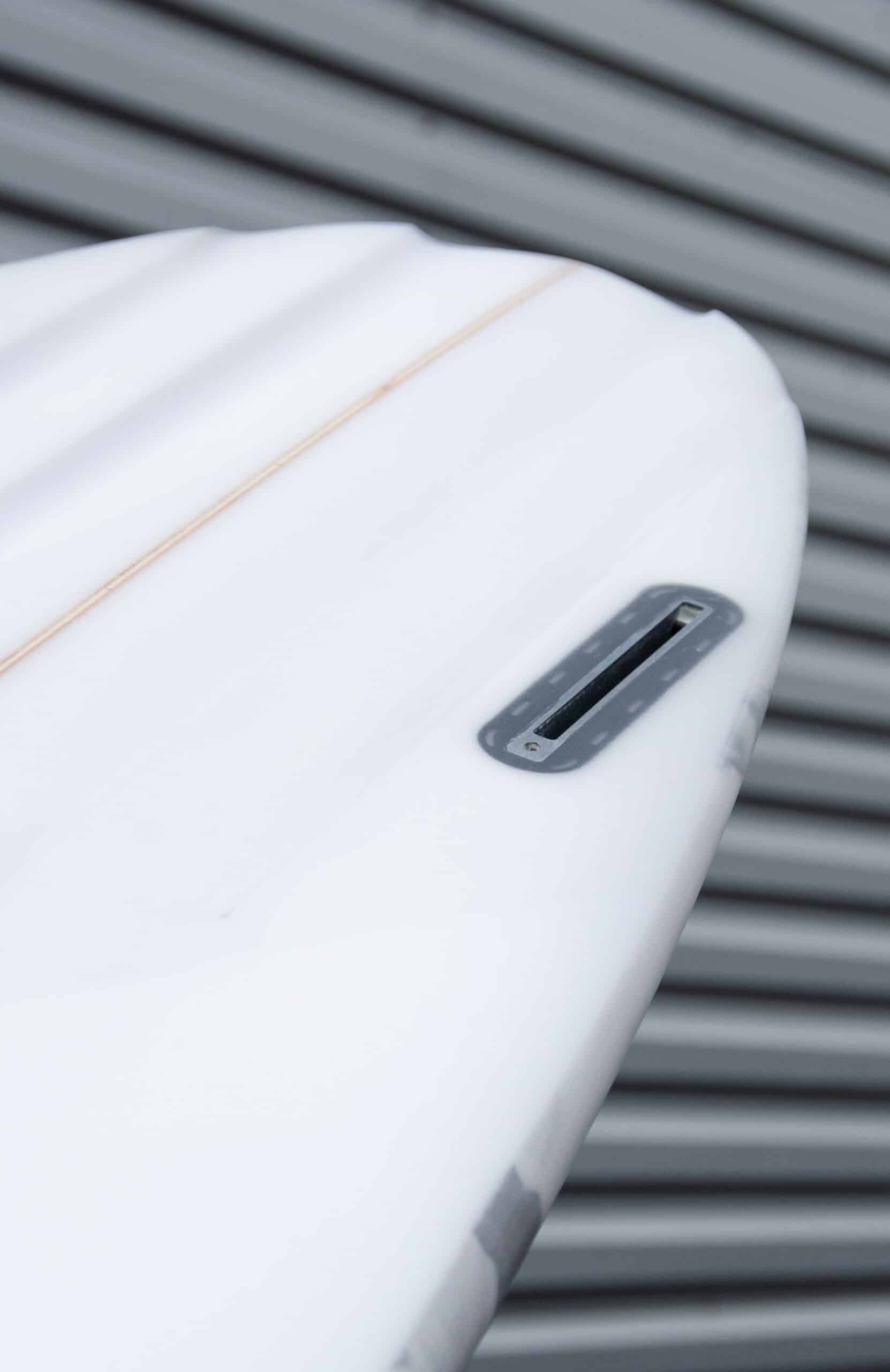 Shapers-Club- Gros plan de la queue d'une planche de surf Aphex 7'0" aux lignes épurées sur un fond flou de lignes grises horizontales. Le boîtier de dérive est visible et la planche présente une surface lisse avec une légère ligne orange qui la longe, rappelant le style Aphex. -surfshop-surfboard