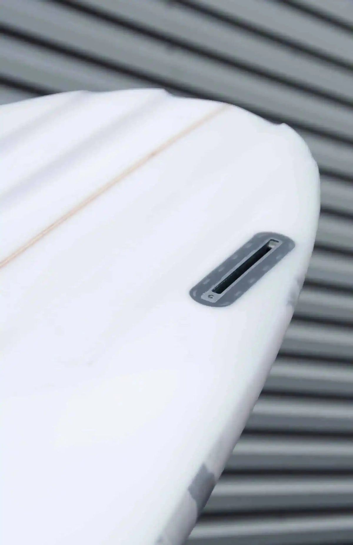 Shapers-Club- Gros plan de la queue d'une planche de surf Aphex 7'0" aux lignes épurées sur un fond flou de lignes grises horizontales. Le boîtier de dérive est visible et la planche présente une surface lisse avec une légère ligne orange qui la longe, rappelant le style Aphex. -surfshop-surfboard