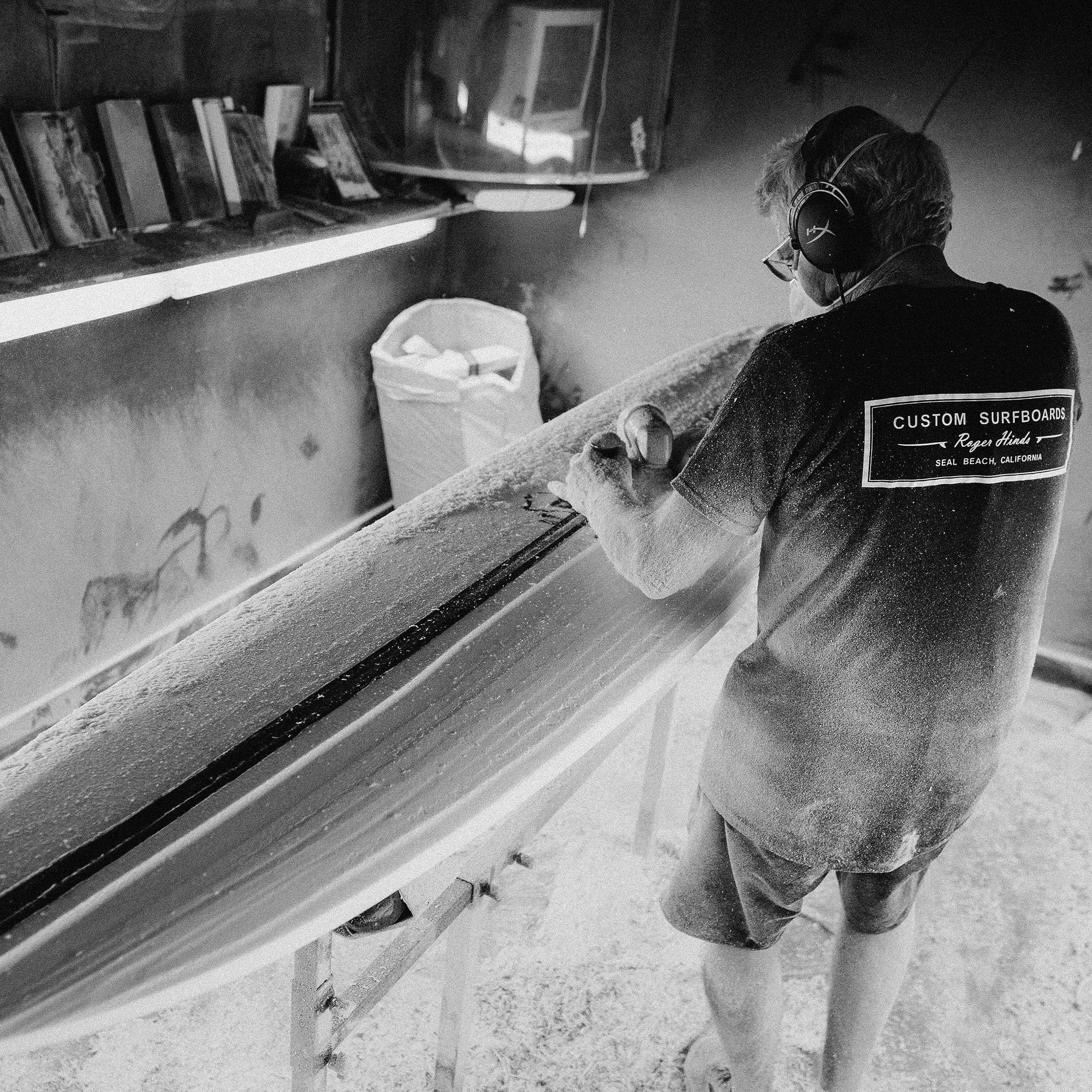 Shapers-Club- Portant un casque et un t-shirt « Custom Surfboards, Seaside, CA », un artisan façonne une planche dans un atelier noir et blanc, un hommage à l'art derrière la planche de surf Acompte deposite - Roger Hinds. -surfshop-surfboard