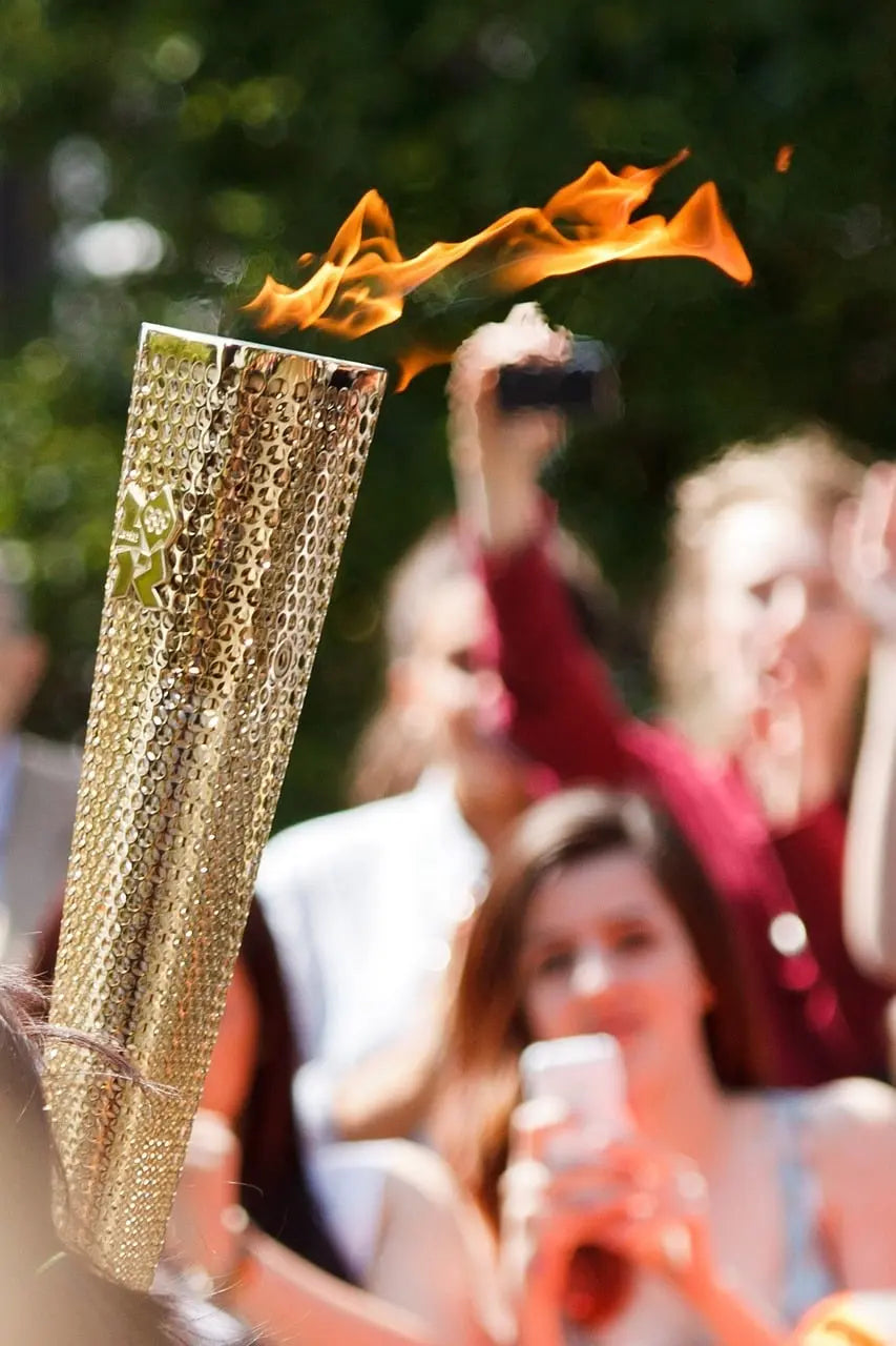 Shapers-Club- Une torche avec une flamme flamboyante est brandie au premier plan, symbolisant Paris 2024, avec des spectateurs en arrière-plan flou, certains capturant l'instant sur leur smartphone. -surfshop-surfboard