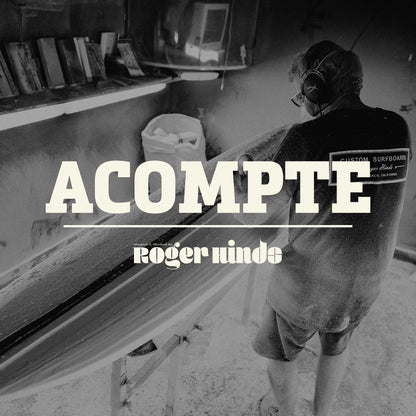 Shapers-Club- Dans un atelier faiblement éclairé, un individu masqué et portant un casque audio façonne minutieusement une planche de surf. Les mots « Acompte deposite - Roger Hinds » sont bien visibles, soulignant le travail artisanal en cours. -surfshop-surfboard