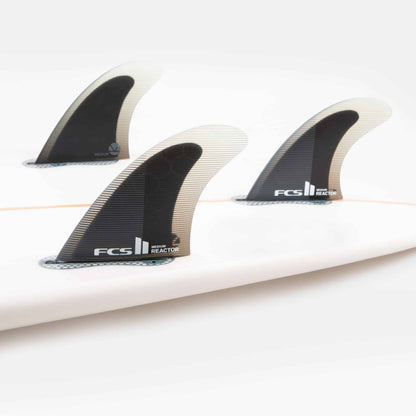 Shapers-Club- Gros plan de trois ailerons de planche de surf noirs et blancs installés sur une planche de surf blanche. Les ailerons FCS II - Reactor - Thruster (Performance Core) portent le texte « FCS II Reactor ». L'arrière-plan est blanc uni, mettant en valeur les ailerons et le design élégant de la planche de surf. -surfshop-surfboard