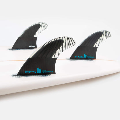 Shapers-Club- Image en gros plan d'une planche de surf blanche équipée de trois ailerons noirs FCS II - PERFORMANCE - THRUSTER (Performance Core Carbon) avec un logo bleu et le texte « Performer » sur chaque aileron, disposés selon une disposition de type thruster axée sur la performance. Le fond est blanc uni. -surfshop-surfboard