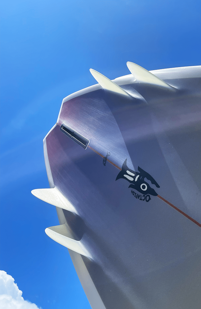 Shapers-Club- Une planche de surf Octafish de 5'10", dotée d'une surface blanche élégante avec plusieurs ailerons, est photographiée d'en bas sur fond de ciel bleu vif. Elle présente un design graphique noir saisissant et porte le logo "02" ainsi que le nom "Octafish", alors qu'elle glisse sous des nuages duveteux au bord de l'image. -surfshop-surfboard