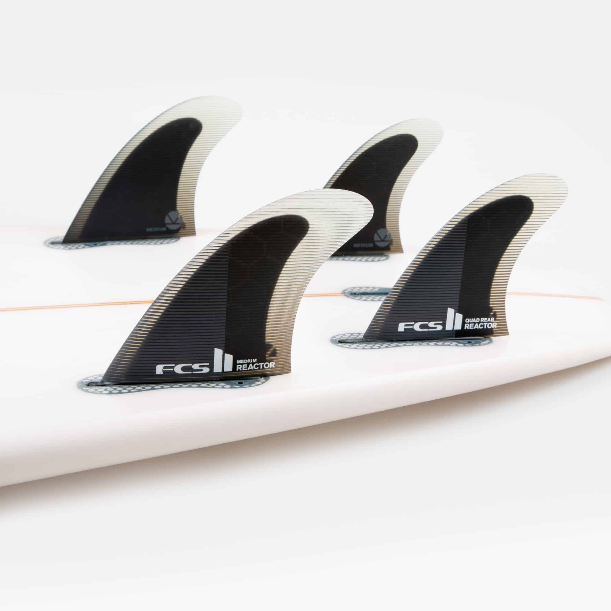 Shapers-Club- Vue rapprochée d'une planche de surf équipée de quatre ailerons FCS II Reactor. Les ailerons QUAD REAR sont noirs avec des bords transparents et étiquetés « FCS II » et « Reactor ». La planche de surf a un design minimaliste et est principalement blanche. -surfshop-surfboard