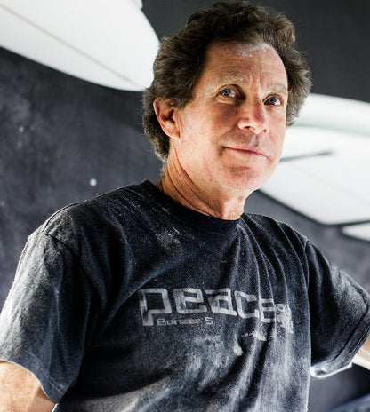 Shapers-Club- Une personne vêtue d'un T-shirt Acompte deposite - Campbell Brothers de couleur sombre avec un motif imprimé fait face à la caméra sur un fond sombre. Sa chemise semble parsemée de taches blanches, tandis que l'éclairage met en valeur son visage et le haut de son corps. -surfshop-surfboard