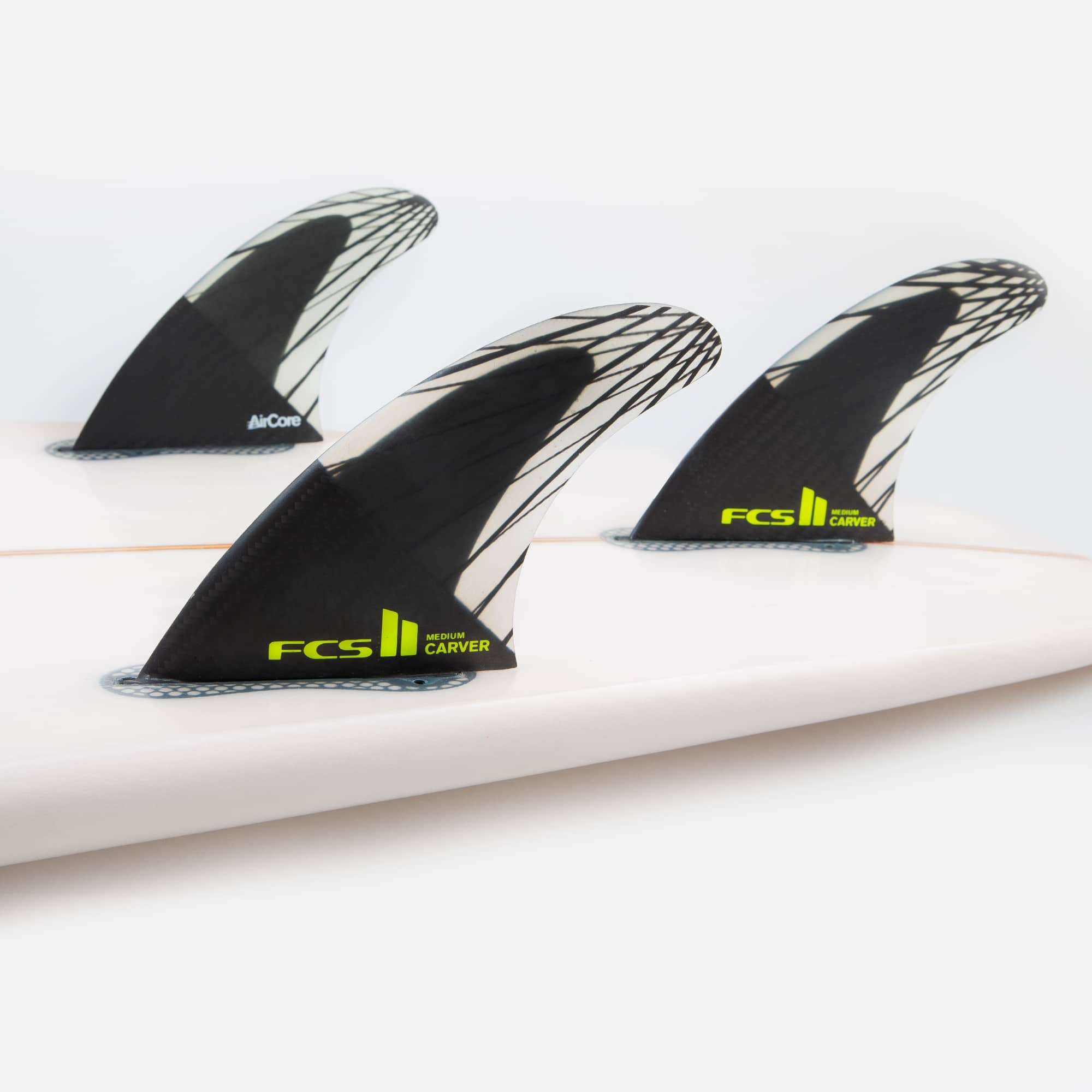 Shapers-Club- Gros plan de trois ailerons de surf FCS II - CARVER - THRUSTER (Performance Core Carbon) noirs et blancs, marqués « Carver » en vert fluo, fixés sur une planche de surf blanche. Les ailerons thruster présentent un motif géométrique distinctif et sont conçus pour améliorer les performances en surf. -surfshop-surfboard
