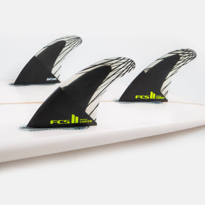 Shapers-Club- Gros plan de trois ailerons de surf FCS II - CARVER - THRUSTER (Performance Core Carbon) noirs et blancs, marqués « Carver » en vert fluo, fixés sur une planche de surf blanche. Les ailerons thruster présentent un motif géométrique distinctif et sont conçus pour améliorer les performances en surf. -surfshop-surfboard