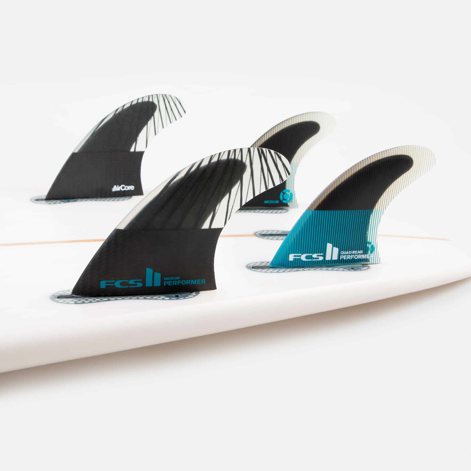 Shapers-Club- Gros plan de trois ailerons de planche de surf fixés sur une planche de surf blanche. Les ailerons FCS II Performer, avec leurs designs élégants en noir, blanc et bleu sarcelle et fabriqués à partir de Performance Core Carbon pour une stabilité exceptionnelle, sont prêts pour la configuration quad ultime. L'arrière-plan est une surface blanche unie. -surfshop-surfboard