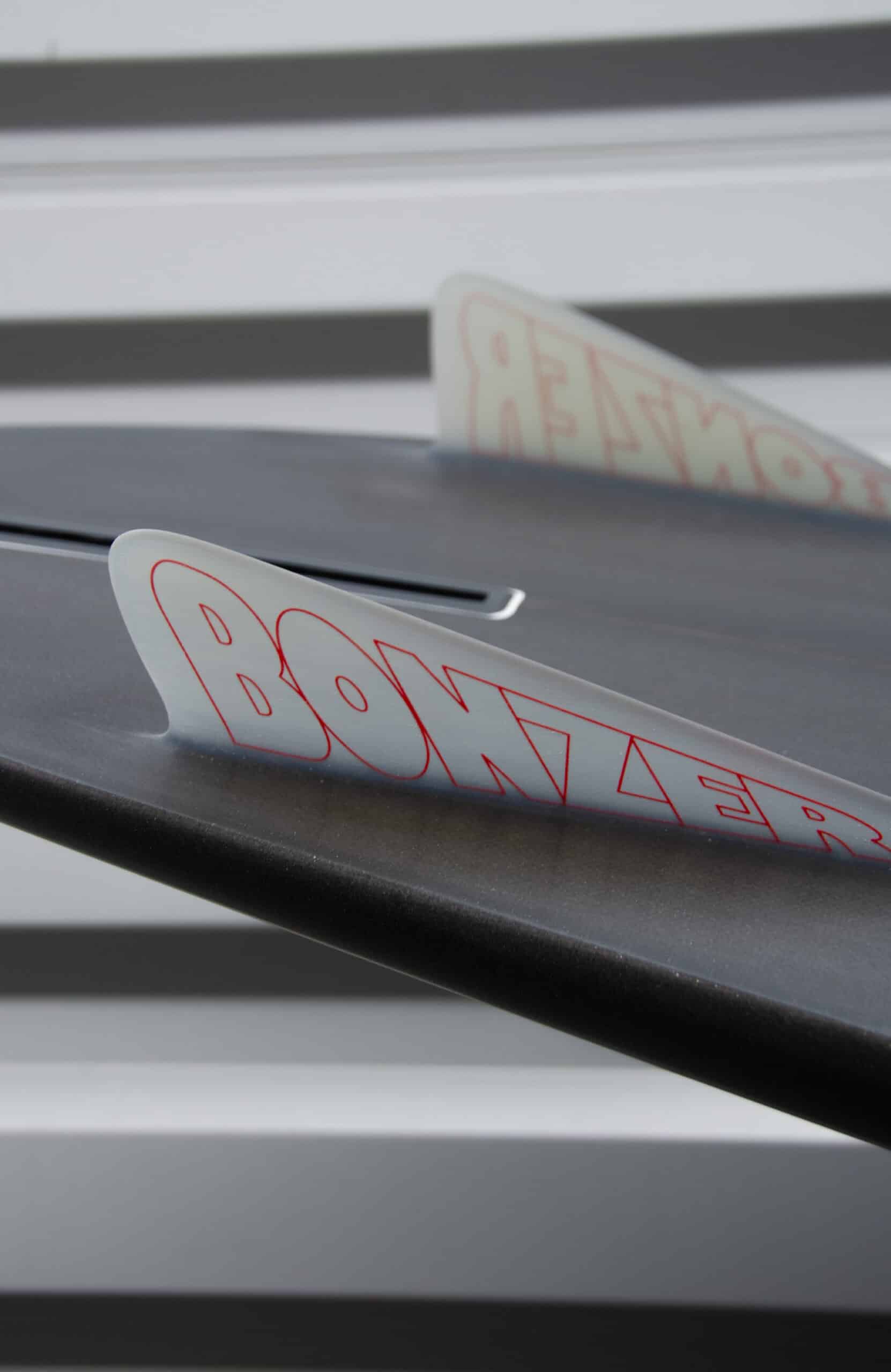 Shapers-Club- Gros plan d'un aileron de planche de surf Campbell Brothers - Russ Short avec le mot « Bonzer » écrit dessus en lettres rouges. L'arrière-plan présente des rayures horizontales floues grises et blanches. L'accent est mis sur l'aileron, offrant une vue partielle du design élégant de la planche de surf de 7'0" x 20' 3/4" x 2' 3/4". -surfshop-surfboard