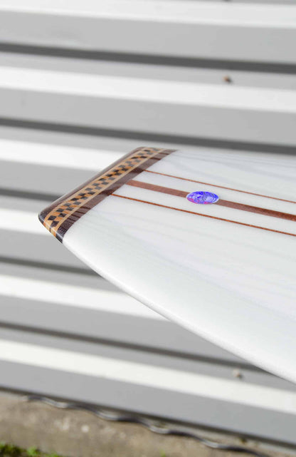 Shapers-Club- Une image en gros plan présente la queue d'une planche de surf Roger Hinds - The Classic contre un mur à rayures horizontales. Mesurant 9'6" x 23" x 3' 3/4", cette planche de surf présente un design élégant avec des incrustations de bois foncé et un petit autocollant circulaire, ajoutant une touche exquise de savoir-faire et de détail à son apparence. -surfshop-surfboard