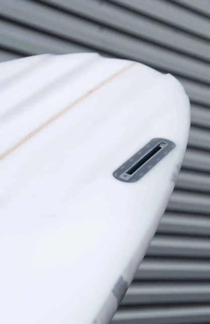 Shapers-Club- Gros plan de la queue d'une planche de surf Aphex 7'0" aux lignes épurées sur un fond flou de lignes grises horizontales. Le boîtier de dérive est visible et la planche présente une surface lisse avec une légère ligne orange qui la longe, rappelant le style Aphex. -surfshop-surfboard