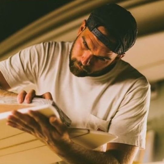 Shapers-Club- Un homme portant un t-shirt blanc et une casquette ponce soigneusement une planche de surf, mettant en valeur le savoir-faire reconnu de DeadKooks. Un éclairage chaleureux accentue sa concentration alors qu'il façonne la planche avec précision, reflétant le style caractéristique qui a fait la renommée d'Eden SAUL dans la collection Acompte deposite. -surfshop-surfboard