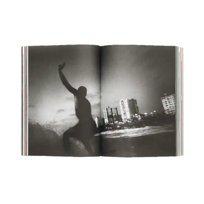 Shapers-Club- An open copy of ACID MAGAZINE - N°6 - WHATEVER SURFING MEANS presents a black and white surf photo: a surfer is surfing a wave near the city skyscrapers under a cloudy sky. -surfshop-surfboard